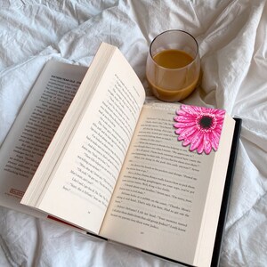 Puede incluir: Un libro abierto con un marcap&aacute;ginas de margarita rosa y un vaso de caf&eacute; sobre una s&aacute;bana blanca. Las p&aacute;ginas del libro son visibles, con texto y un borde rojo. El marcap&aacute;ginas es una flor rosa brillante con un centro negro.
