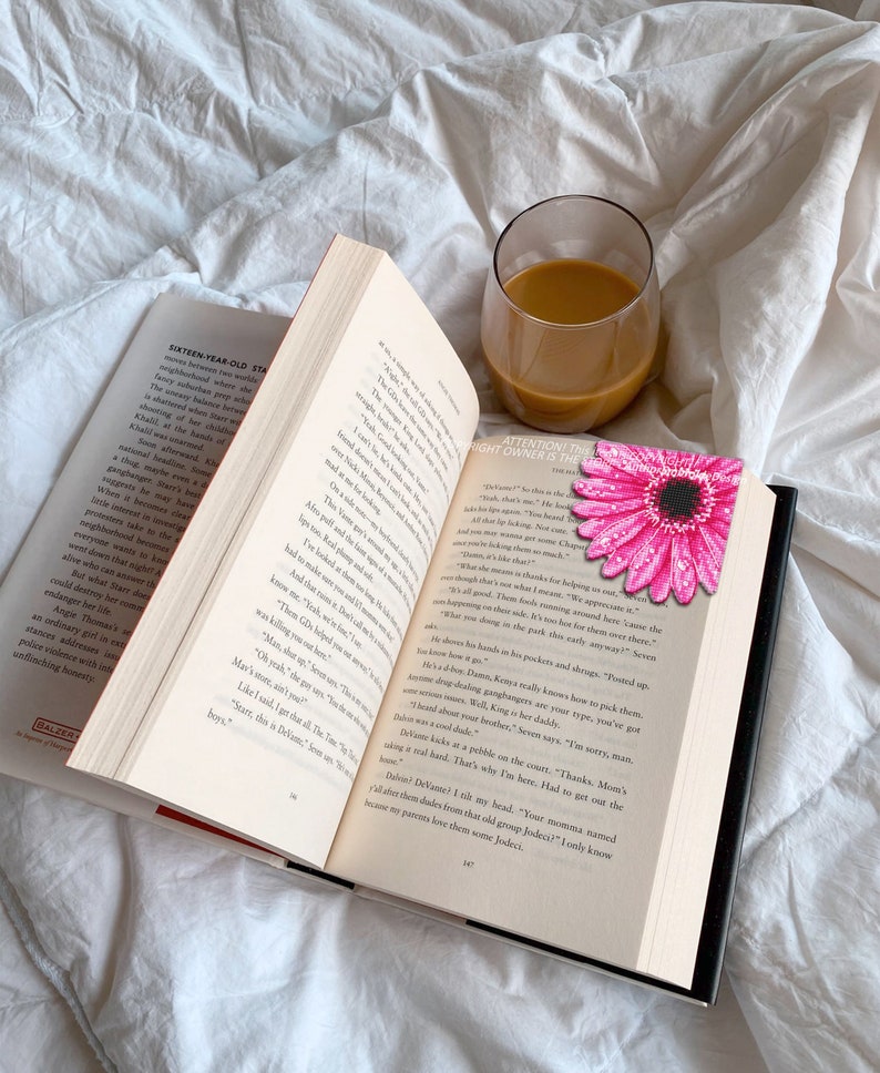 Puede incluir: Un libro abierto con un marcap&aacute;ginas de margarita rosa y un vaso de caf&eacute; sobre una s&aacute;bana blanca. Las p&aacute;ginas del libro son visibles, con texto y un borde rojo. El marcap&aacute;ginas es una flor rosa brillante con un centro negro.