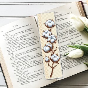 May include: A white cotton bookmark with a brown stem and leaves. The cotton is arranged in a vertical line with the largest cotton boll at the top and the smallest at the bottom.