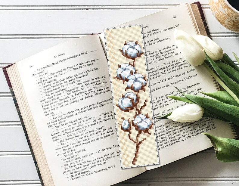 May include: A white cotton bookmark with a brown stem and leaves. The cotton is arranged in a vertical line with the largest cotton boll at the top and the smallest at the bottom.