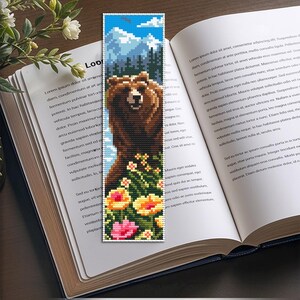 May include: A pixelated bookmark featuring a brown bear standing in front of a mountain range and blue sky. The bear is surrounded by colorful flowers. The bookmark is on top of an open book.