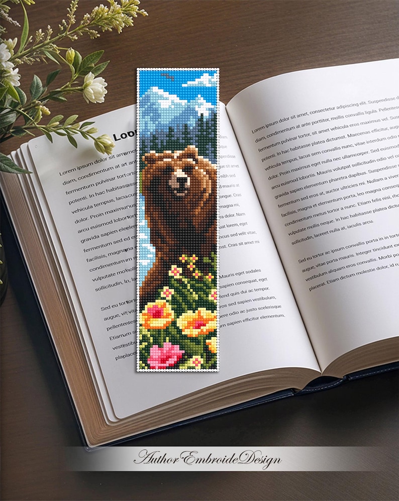 May include: A pixelated bookmark featuring a brown bear standing in front of a mountain range and blue sky. The bear is surrounded by colorful flowers. The bookmark is on top of an open book.