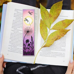 May include: A handmade bookmark featuring a dandelion and dragonfly design. The bookmark has a gradient of pink and purple hues, with a yellow sun and black silhouettes. A book is open with a yellow leaf resting on the pages.