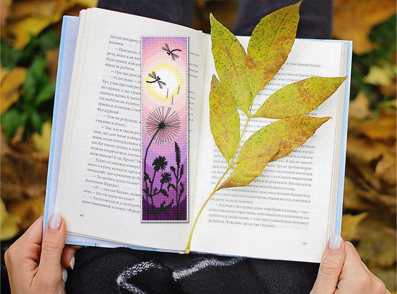May include: A handmade bookmark featuring a dandelion and dragonfly design. The bookmark has a gradient of pink and purple hues, with a yellow sun and black silhouettes. A book is open with a yellow leaf resting on the pages.