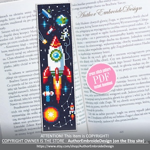 May include: A cross-stitch bookmark featuring a colorful rocket ship blasting off into space, surrounded by planets, stars, and an astronaut. The bookmark is set against the backdrop of an open book.