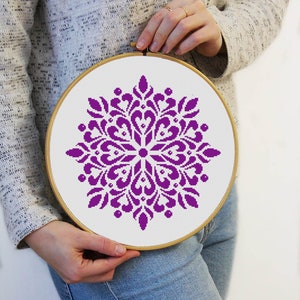 May include: A round embroidery hoop with a white fabric featuring a purple floral cross-stitch design. The design is a symmetrical pattern with heart-shaped and petal-like elements. The hoop is held by a person.