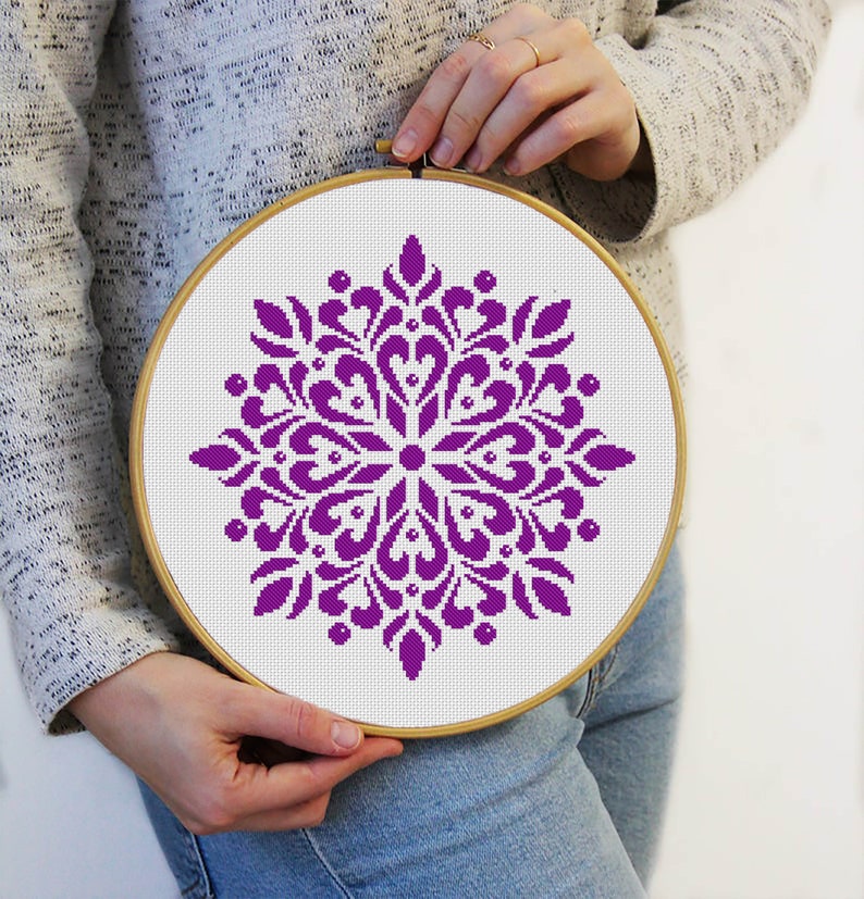 May include: A round embroidery hoop with a white fabric featuring a purple floral cross-stitch design. The design is a symmetrical pattern with heart-shaped and petal-like elements. The hoop is held by a person.