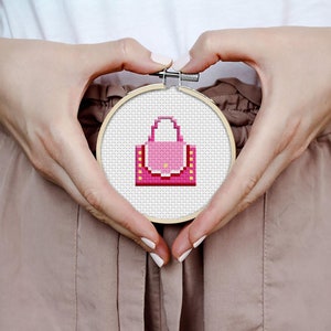 May include: A pink and yellow cross-stitch embroidery of a handbag in a wooden embroidery hoop.
