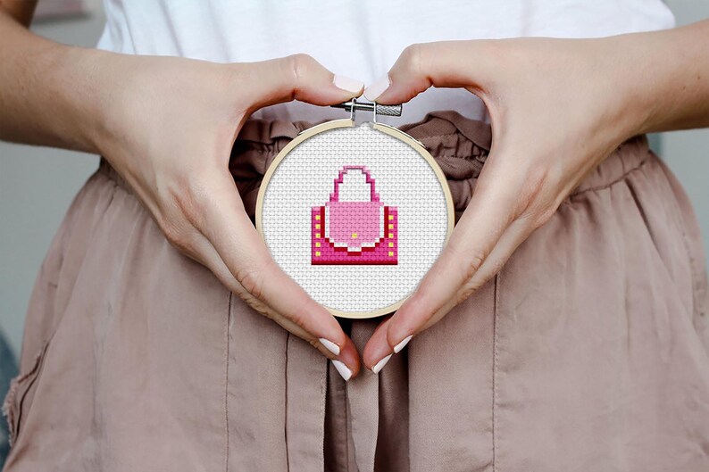 May include: A pink and yellow cross-stitch embroidery of a handbag in a wooden embroidery hoop.