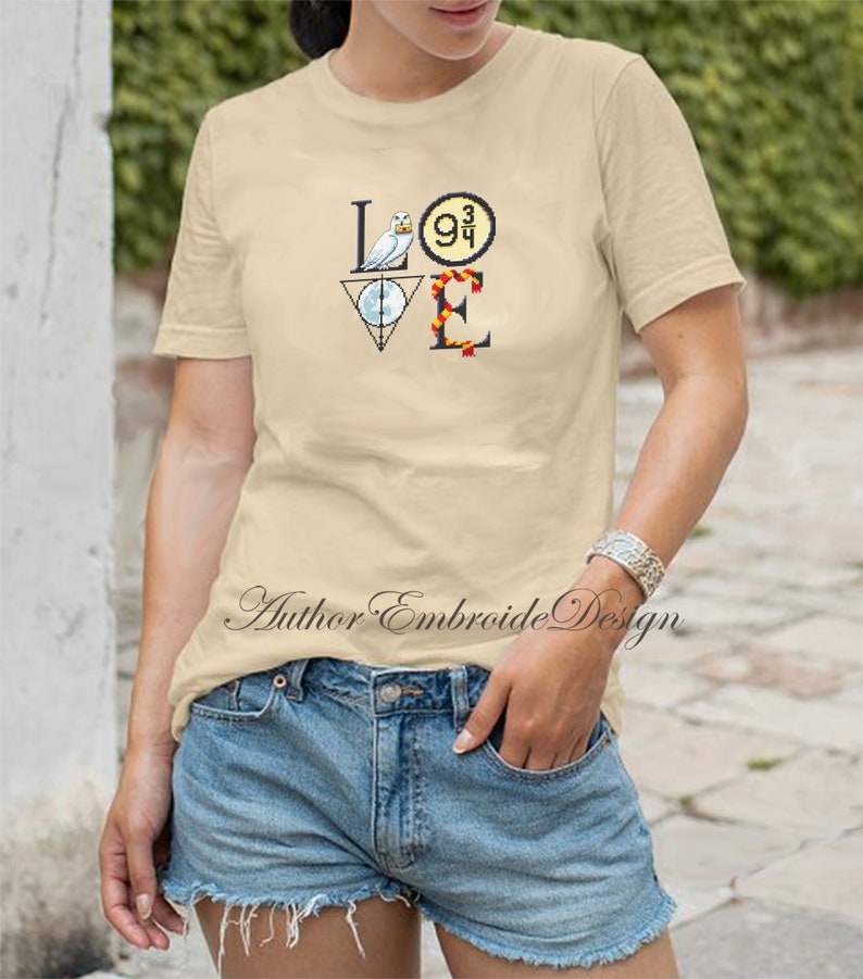 May include: A beige t-shirt with a graphic design of the word "LOVE" made up of Harry Potter themed icons. The "L" is a white owl, the "O" is a golden snitch, the "V" is a triangle with a circle inside, and the "E" is a red and gold scarf.