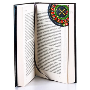 Op de afbeelding: Een close-up van een boek met een bladwijzer met een kleurrijk roulettewiel met een rode X in het midden. De bladwijzer is gemaakt van stof en heeft een zwart lint.