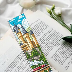 Puede incluir: Un marcap&aacute;ginas hecho a mano con una imagen pixelada de un castillo con altas torres, un tren y un puente. El marcap&aacute;ginas est&aacute; colocado sobre un libro abierto y una vela encendida. El texto "Author Embroide Design" es visible.
