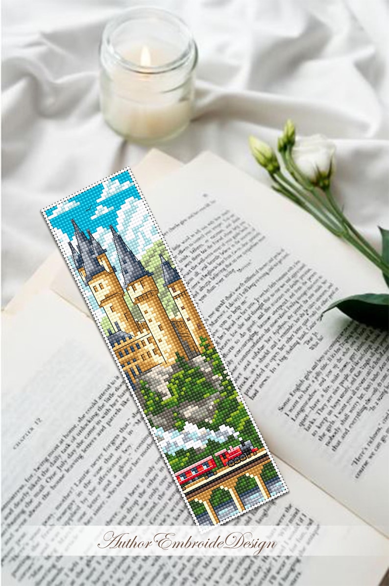 Puede incluir: Un marcap&aacute;ginas hecho a mano con una imagen pixelada de un castillo con altas torres, un tren y un puente. El marcap&aacute;ginas est&aacute; colocado sobre un libro abierto y una vela encendida. El texto "Author Embroide Design" es visible.