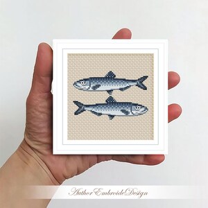 May include: A cross-stitch embroidery design featuring two blue and white fish on a beige background. The design is framed in a white frame.