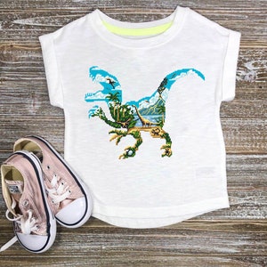 May include: White t-shirt with a colorful dinosaur graphic. The dinosaur is made up of a jungle scene with palm trees, mountains, and a blue sky.