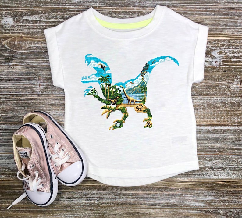 May include: White t-shirt with a colorful dinosaur graphic. The dinosaur is made up of a jungle scene with palm trees, mountains, and a blue sky.