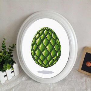 May include: A framed cross-stitch of a green, scaled egg. The egg is centered within a white oval frame. The cross-stitch design features varying shades of green, creating a textured appearance. The frame is set against a white background.