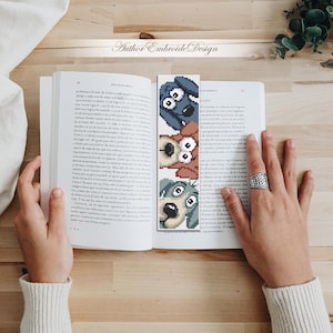 May include: A handmade bookmark with three cartoon dog faces in blue, brown, and green. The bookmark is in an open book on a wooden surface. The text "Author Embroide Design" is visible.