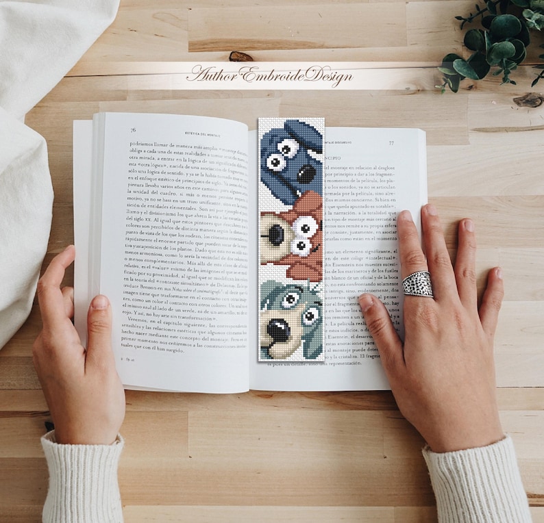 May include: A handmade bookmark with three cartoon dog faces in blue, brown, and green. The bookmark is in an open book on a wooden surface. The text "Author Embroide Design" is visible.