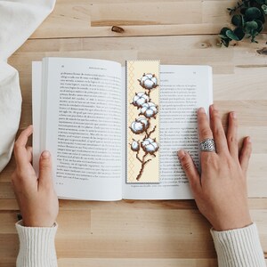 May include: A beige bookmark featuring a cross-stitch design of a cotton plant with white cotton balls. The plant has a brown stem and green leaves.