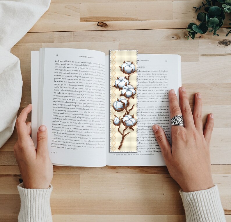 May include: A beige bookmark featuring a cross-stitch design of a cotton plant with white cotton balls. The plant has a brown stem and green leaves.