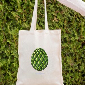 May include: A cream-colored tote bag with a green, pixelated egg design. The egg is a vibrant green with a textured, geometric pattern. The bag is held by a person in front of a green hedge.