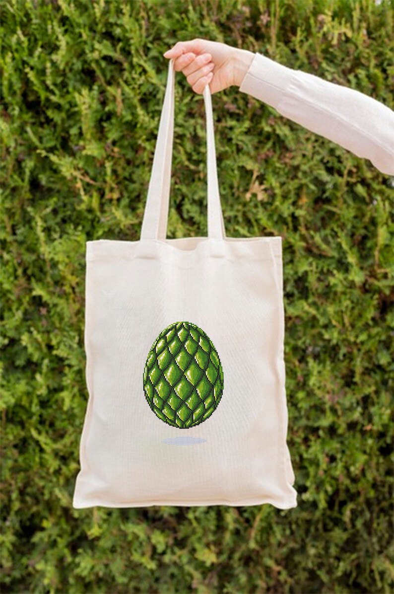 May include: A cream-colored tote bag with a green, pixelated egg design. The egg is a vibrant green with a textured, geometric pattern. The bag is held by a person in front of a green hedge.