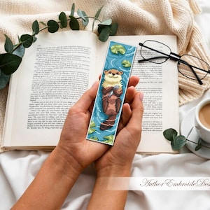 May include: A handmade bookmark featuring a cross-stitch design of an otter floating in water. The bookmark is held in a person's hands, with a book, glasses, and a cup of coffee in the background. The bookmark is approximately 2 inches wide and 6 inches long.