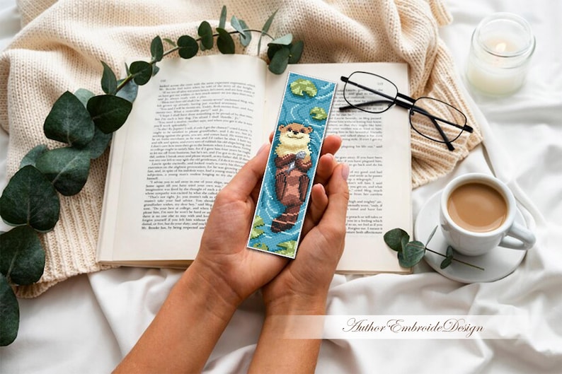 May include: A handmade bookmark featuring a cross-stitch design of an otter floating in water. The bookmark is held in a person's hands, with a book, glasses, and a cup of coffee in the background. The bookmark is approximately 2 inches wide and 6 inches long.
