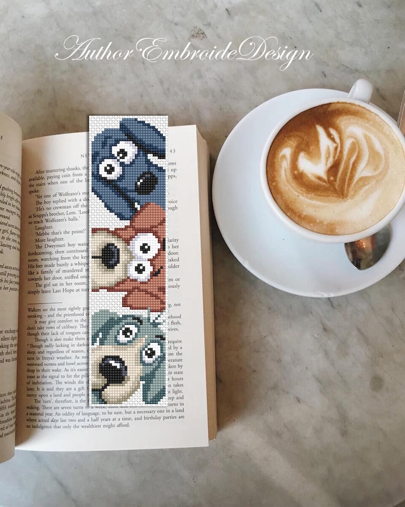May include: An embroidered bookmark with three dog faces in blue, brown, and green, placed in an open book. A cup of coffee with latte art sits beside the book. The text "Author Embroide Design" is visible at the top of the image.