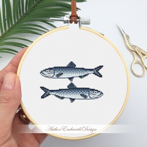 May include: A white embroidery hoop with two blue and gray fish stitched on it. The hoop is held by a hand. The text "Author Embroide Design" is printed on the hoop.