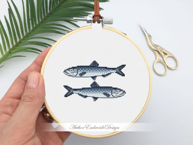 May include: A white embroidery hoop with two blue and gray fish stitched on it. The hoop is held by a hand. The text "Author Embroide Design" is printed on the hoop.