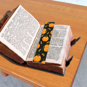 May include: A close-up of a book with a bookmark featuring a pattern of orange pumpkins and green leaves on a black background. The bookmark is tucked into the pages of the book.