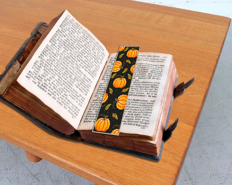 May include: A close-up of a book with a bookmark featuring a pattern of orange pumpkins and green leaves on a black background. The bookmark is tucked into the pages of the book.