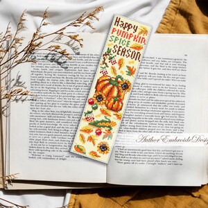 May include: A cross-stitched bookmark with a fall theme, featuring a pumpkin, sunflowers, and the text "Happy Pumpkin Spice Season." The bookmark is placed in an open book.