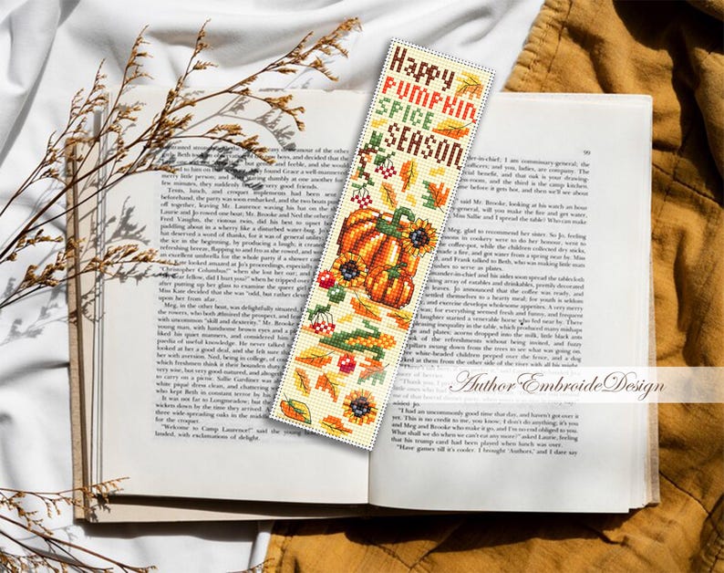 May include: A cross-stitched bookmark with a fall theme, featuring a pumpkin, sunflowers, and the text "Happy Pumpkin Spice Season." The bookmark is placed in an open book.