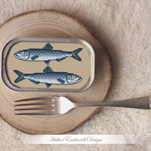 May include: A cross-stitch embroidery design of two sardines in a silver can. The can is on a wooden surface.