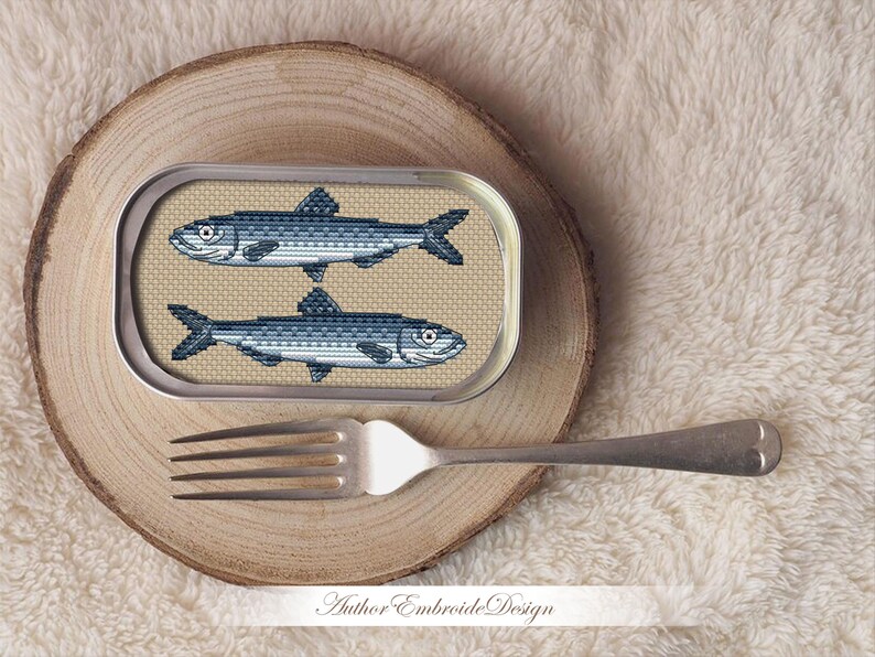 May include: A cross-stitch embroidery design of two sardines in a silver can. The can is on a wooden surface.