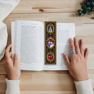 May include: A black and gold bookmark with three images. The top image is a tombstone with the word "RIP" on it. The middle image is a couple of skeletons holding hands. The bottom image is a red heart with a black bat.