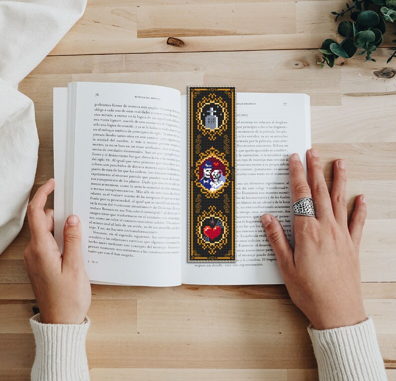 May include: A black and gold bookmark with three images. The top image is a tombstone with the word "RIP" on it. The middle image is a couple of skeletons holding hands. The bottom image is a red heart with a black bat.