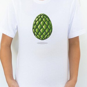 May include: White t-shirt featuring a pixelated green egg design. The egg has a textured, scaled appearance and is centered on the shirt. The shirt is plain and has short sleeves.