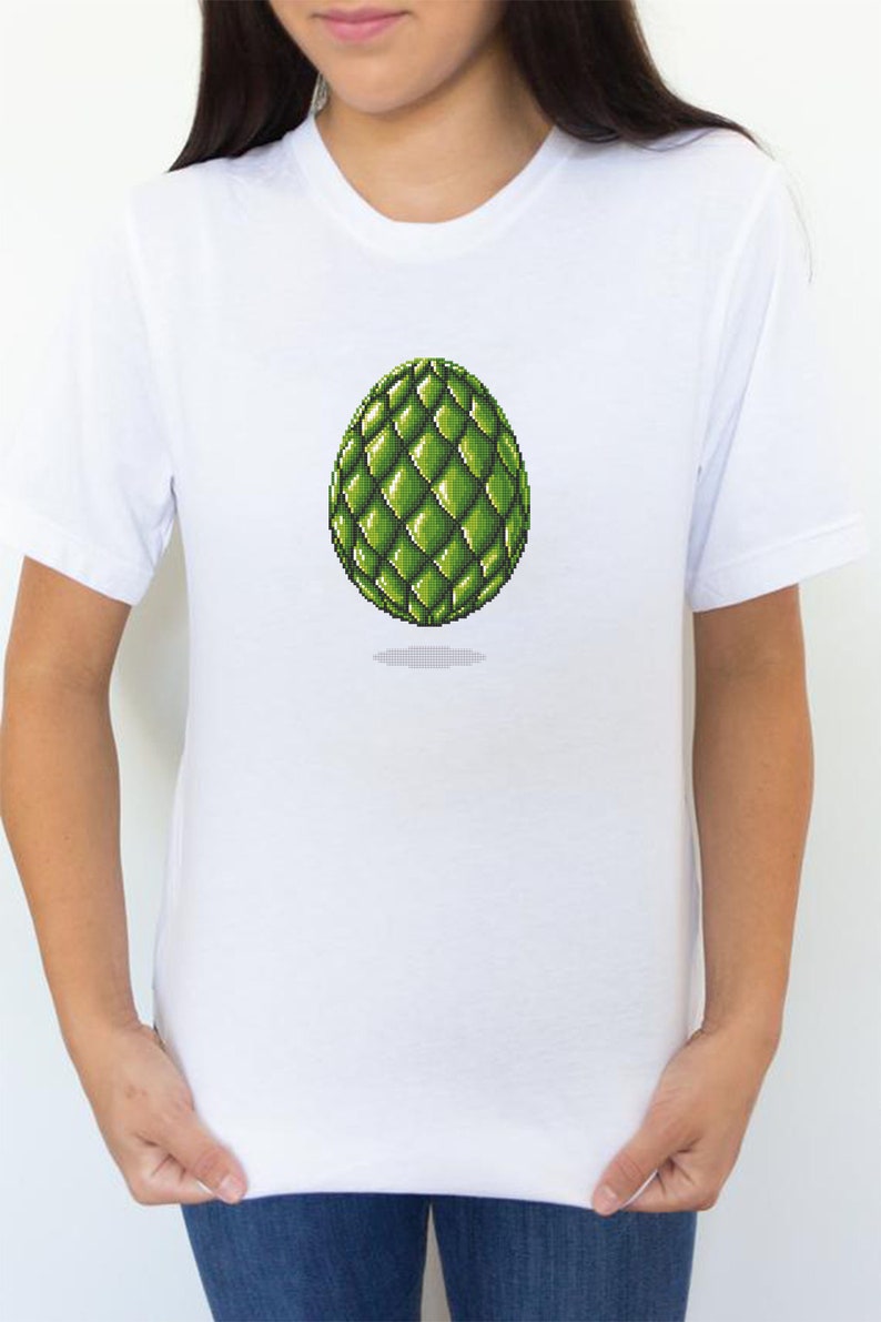 May include: White t-shirt featuring a pixelated green egg design. The egg has a textured, scaled appearance and is centered on the shirt. The shirt is plain and has short sleeves.