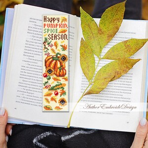 May include: A handmade bookmark with a fall theme, featuring pumpkins, sunflowers, and the text "Happy Pumpkin Spice Season." The bookmark is placed in an open book, with a yellow leaf resting on the pages.