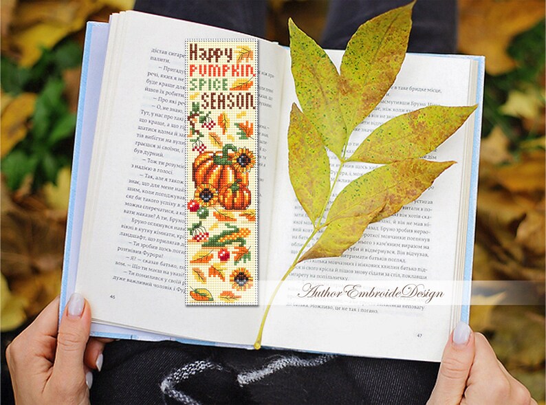 May include: A handmade bookmark with a fall theme, featuring pumpkins, sunflowers, and the text "Happy Pumpkin Spice Season." The bookmark is placed in an open book, with a yellow leaf resting on the pages.