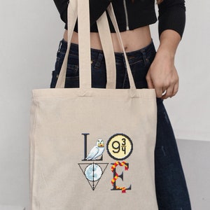 May include: A white canvas tote bag with a black and white graphic of the word "LOVE" with Harry Potter themed icons. The "L" is a white owl, the "O" is a platform 9 3/4 sign, the "V" is a Deathly Hallows symbol, and the "E" is a Gryffindor scarf.