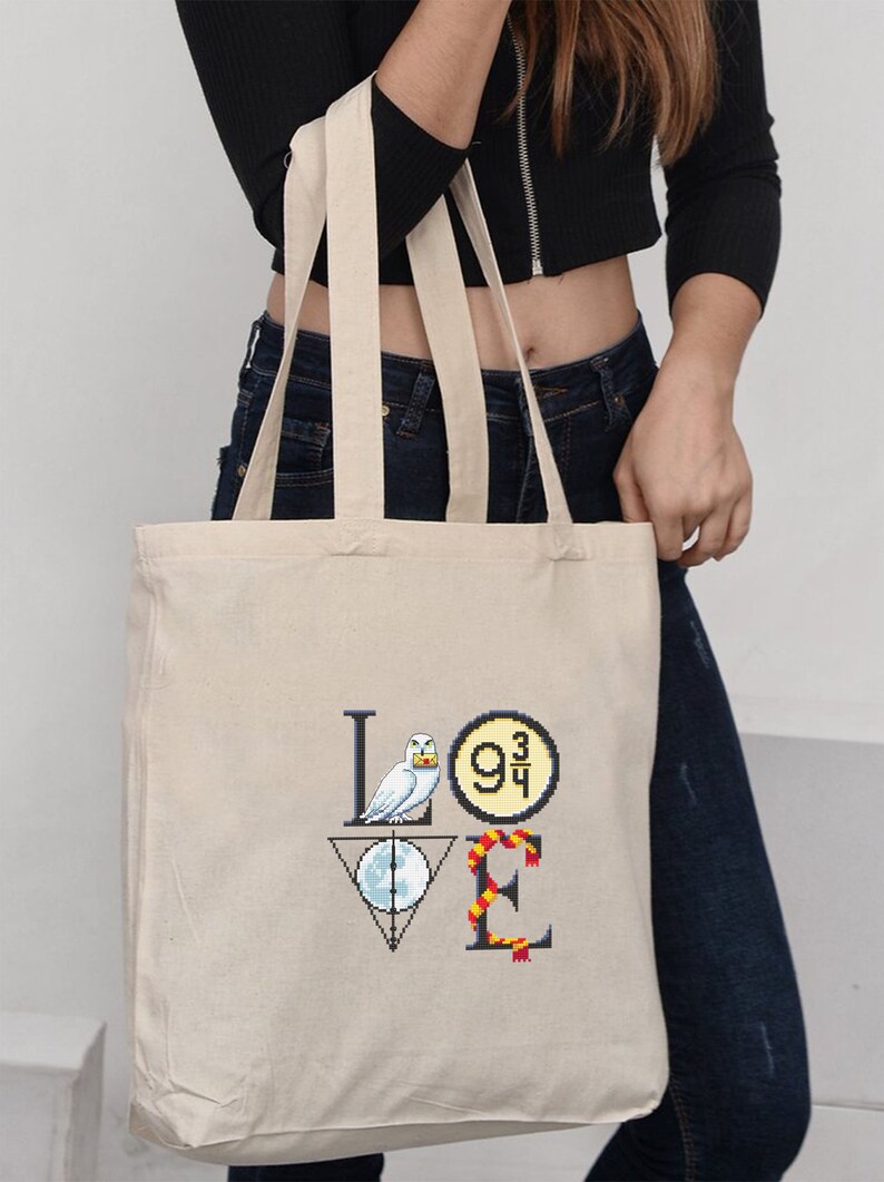 May include: A white canvas tote bag with a black and white graphic of the word "LOVE" with Harry Potter themed icons. The "L" is a white owl, the "O" is a platform 9 3/4 sign, the "V" is a Deathly Hallows symbol, and the "E" is a Gryffindor scarf.