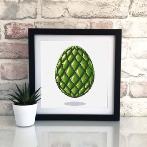 May include: A framed cross-stitch artwork featuring a green, textured egg design. The egg has a diamond pattern and is set against a white background. The frame is black, and a small potted plant is visible to the left.