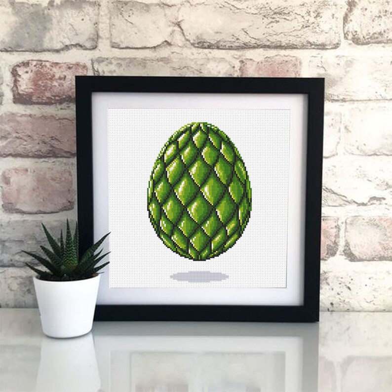 May include: A framed cross-stitch artwork featuring a green, textured egg design. The egg has a diamond pattern and is set against a white background. The frame is black, and a small potted plant is visible to the left.
