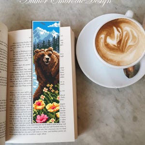 May include: A handmade bookmark featuring a pixelated bear design with a mountain and sky background. The bookmark also includes colorful flowers at the bottom. The bookmark is placed in an open book next to a cup of coffee.