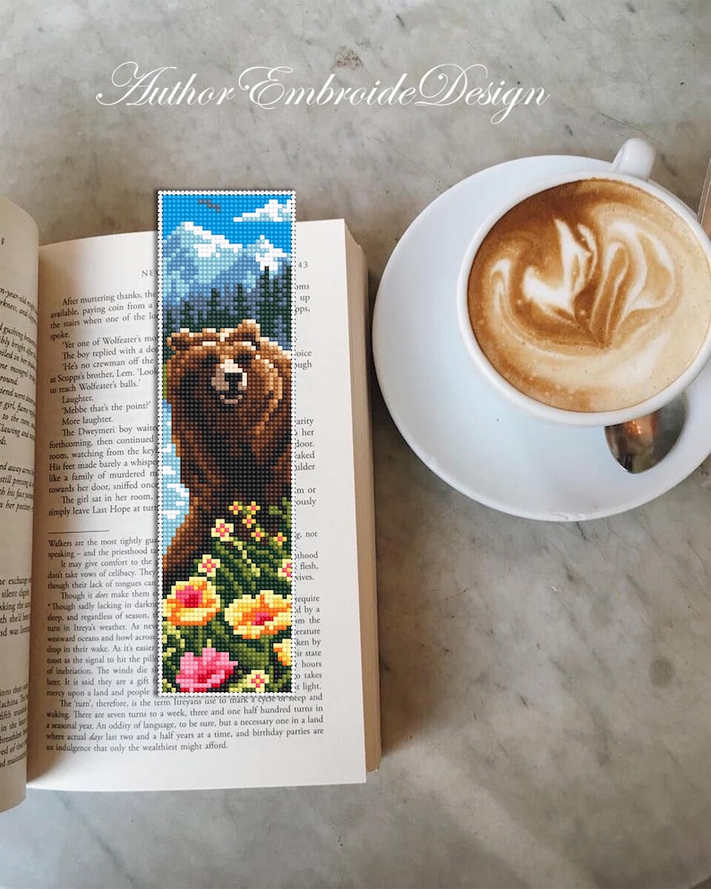 May include: A handmade bookmark featuring a pixelated bear design with a mountain and sky background. The bookmark also includes colorful flowers at the bottom. The bookmark is placed in an open book next to a cup of coffee.
